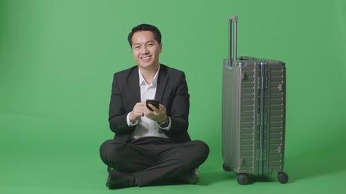 Businessman With Luggage Using Smartphone Then Smiling To Camera While Sitting In The Green Screen