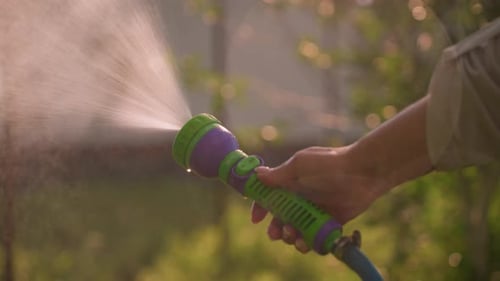 CloseUp of Hand Holding Water Hose Spraying Outdoors with Blurred Greenery Background