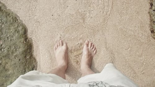 Top view of barefoot male person standing in clear ocean water with sandy ground. Waves crashing and