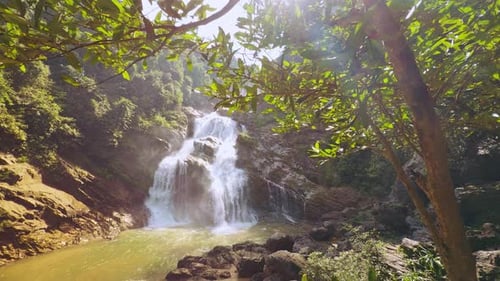 Breiter tropischer Wasserfall, umgeben von dichtem Wald und Ästen mit sanften Sonnenstrahlen und aufgehendem Himmel
