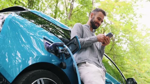 Man stands with phone near her electric car and waits when vehicle will charged at green forest. Sav