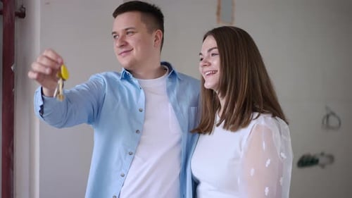 Loving Young Caucasian Couple Standing in New Flat Under Construction Stretching Apartment Key