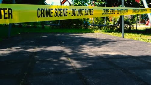 Yellow Crime Scene Do Not Enter tape in front of moving swings at a childrens playground, sunny day.