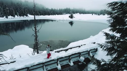 Drone Flying Over Man Snowshoeing Across Bridge With Scenic Alpine Lake Background