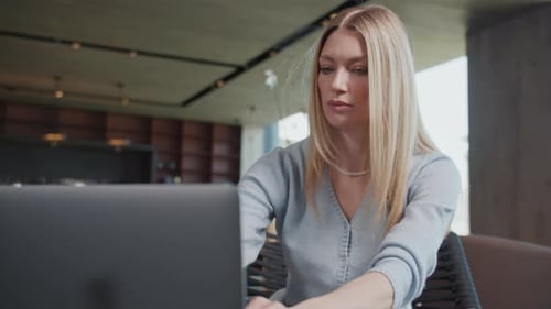 Beautiful Blond Young Woman Using and Typing Laptop in a Cafe Sitting Serious Face Working Freelance