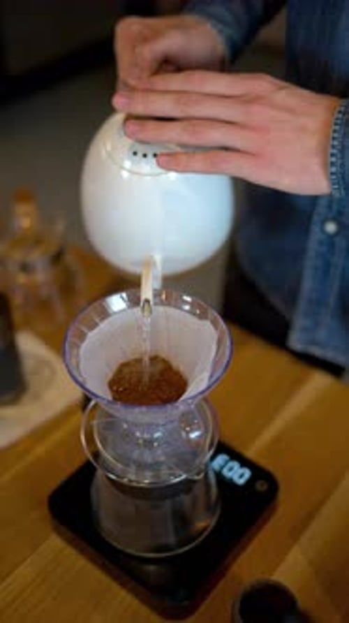 Close up of a man brewing pour-over drip coffee in a slow, circular motion on a wooden table. Vertic