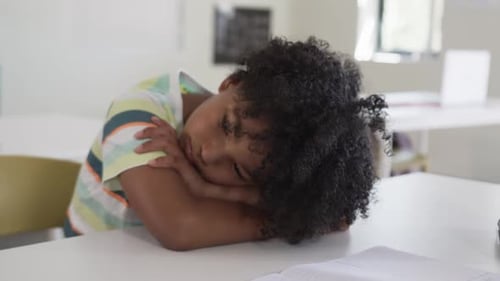 African American child resting on arms at classroom table, glancing papers, pencil after studying
