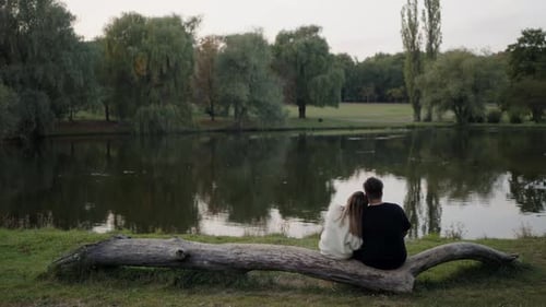 Back view on love couple man and woman sitting cuddling on tree trunk near lake