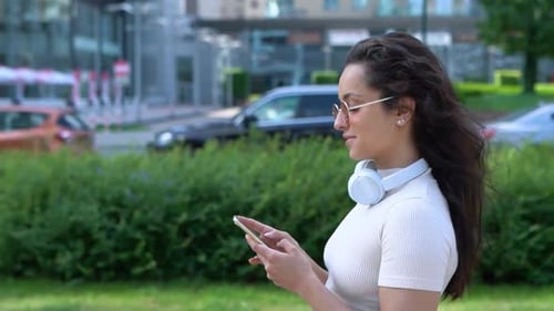 Smiling Young Woman Using Smartphone Typing Message While Walking Along the Street