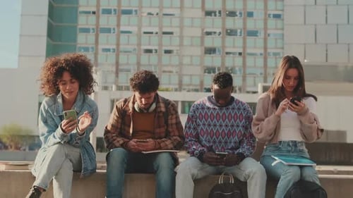 Group of students sit next to the university looking at their mobile phones