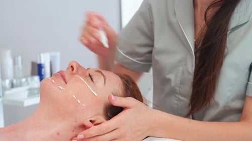 Cosmetologist Applies Cream To Woman's Face At Clinic