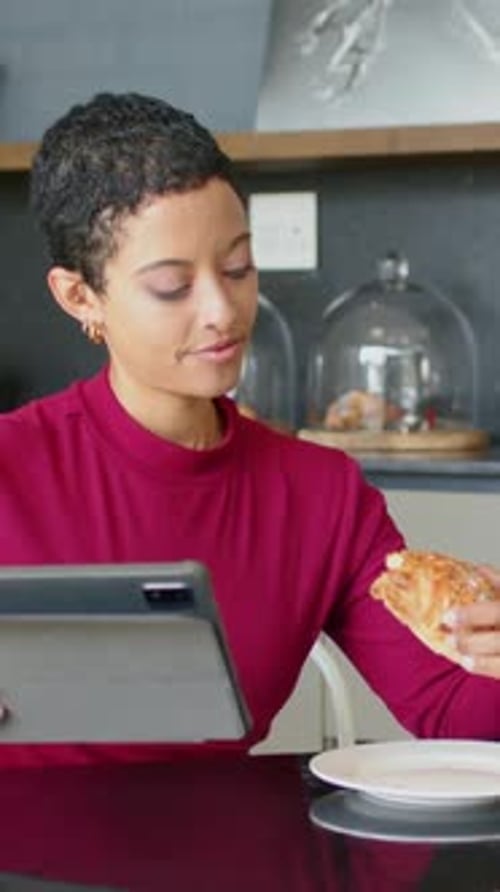 Vertical video: Reading tablet, woman picking and eating pastry at kitchen table in burgundy top