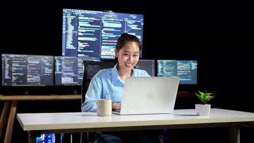 Asian Female Programmer Writing Code By A Laptop Using Multiple Monitors