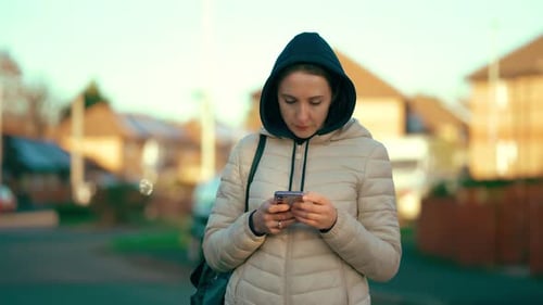 Woman Wearing Hoodie Focused on Smartphone While Standing in Quiet Residential Street Female