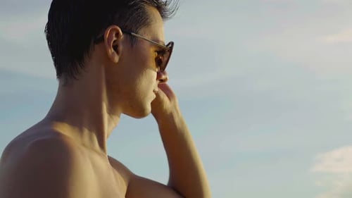 Homem caucasiano sem camisa usando óculos escuros olha para o mar, vista clara do céu