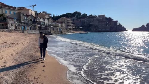 A Woman Walks Along the Sandy Seashore Against the Backdrop of a Resort Town