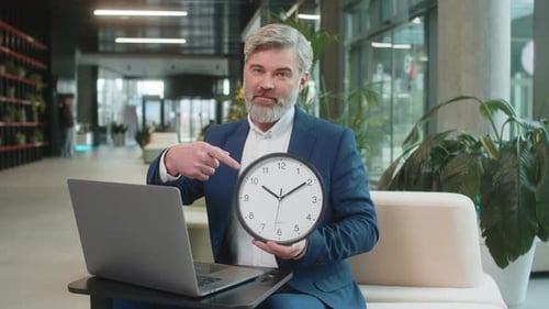 Mature Businessman with Laptop in Lobby Showing Time on Wall Clock and Pointing the Finger at Camera