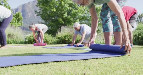 Diverse group of male and female seniors rolling up yoga mats after exercise in garden, slow motion
