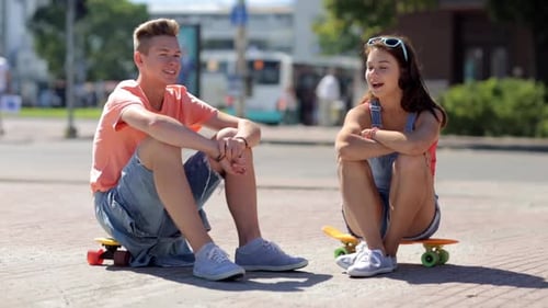 Smiling teenage couple chatting on city street with skateboards in summer