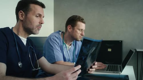Side view of two male caucasian doctors discussing over medical X-ray scan on board and laptop. Shot