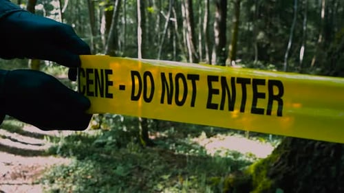 Crime Scene tape being unraveled and pulled infront of a forest hiking road by investigator hands.