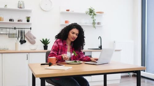 Woman Eats Salad While Using Laptop in Kitchen
