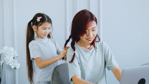 Child Braids Woman's Hair While She Works on Laptop