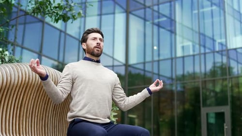 Businessman practicing meditation sitting on bench outside modern glass building.