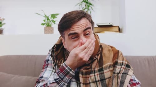 Un homme âgé enveloppé dans une couverture douillette éternuant, toussant et luttant contre un rhume dans un endroit lumineux