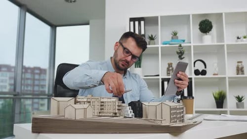 Man engineer which working with constructed model of new building checking datas from in design offi