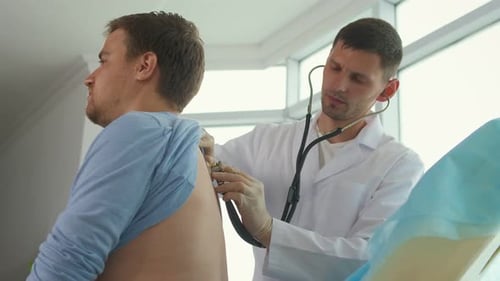 A Doctor Listens to Lungs with a Stethoscope To a Young Man in a Clinic Office