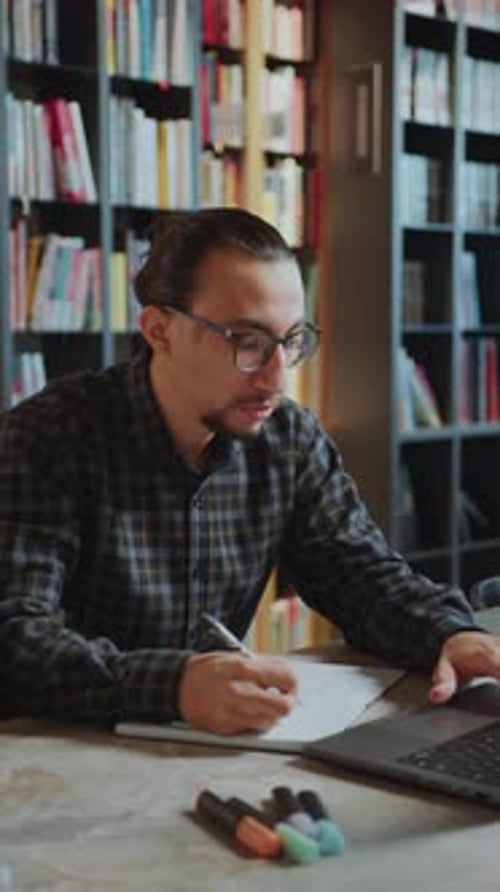 Male Student Using Laptop and Writing in Copybook in Library