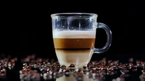 Closeup Latte in Glass Cup with Roasted Coffee Beans Falling in Slow Motion on Table Closeup Tasty