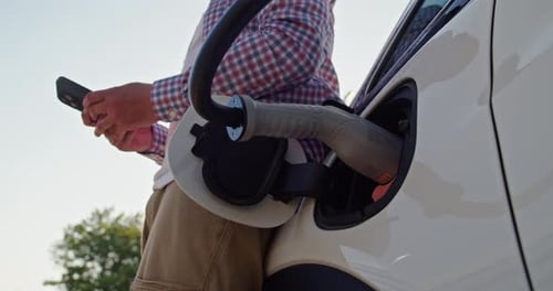 Man With Mobile Phone Stands Near Electric Car Connected to a Charging Station
