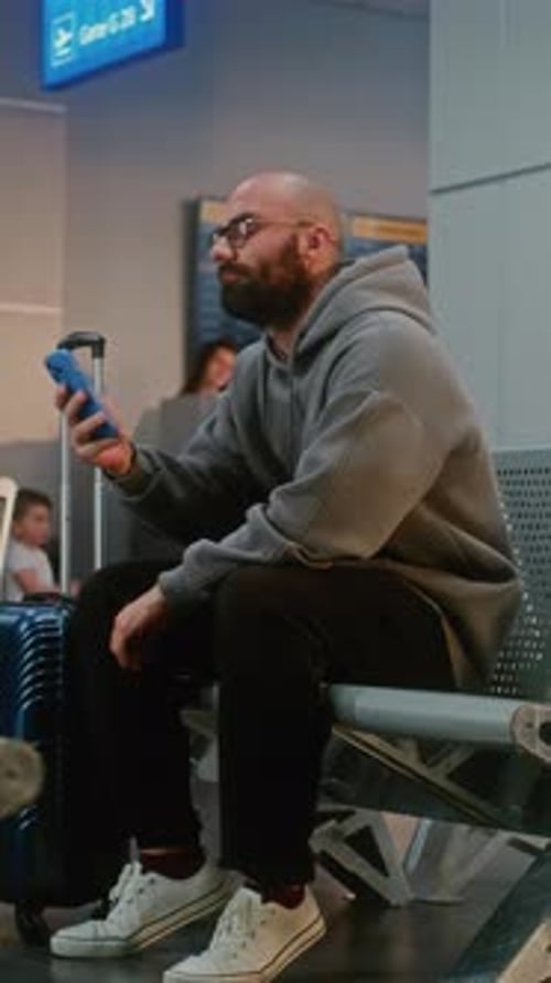 Airport Terminal Man with Suitcase Sitting in Departure Lounge After Security Check Using Phone