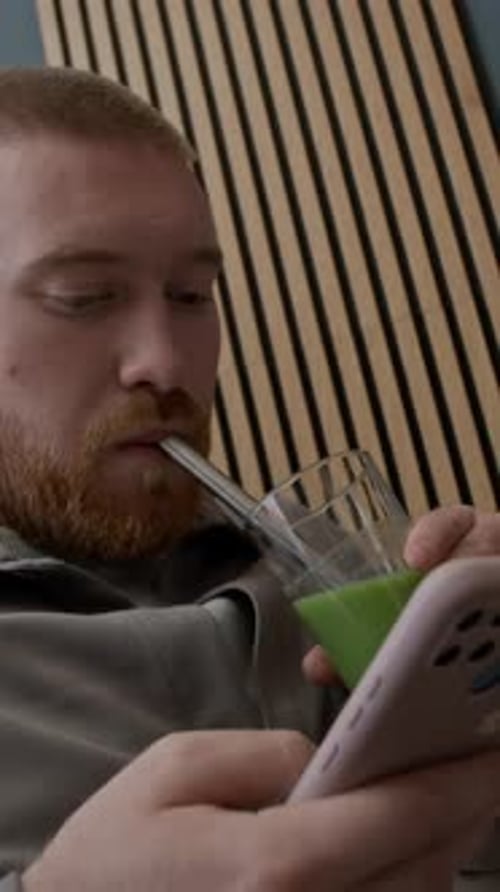 Vertical of Young Man Drinking Smoothie with Straw while Scrolling Through Phone