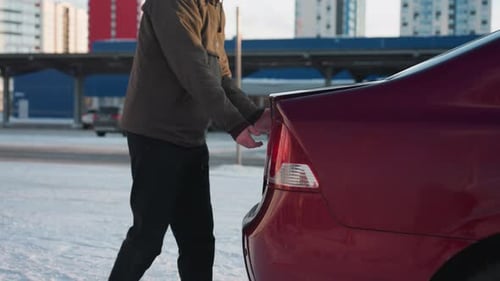 Man Approaches Parked Red Car and Opens Rear Trunk in Snowy Lot