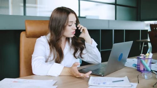 Businesswoman Engaged in Phone Conversation While Working on Laptop in Modern Office Space During