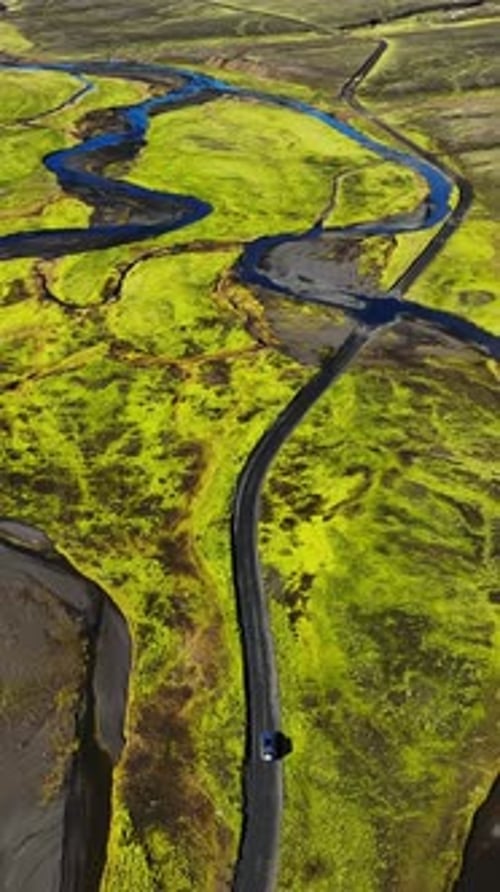 Vertical drone shot of a lone SUV driving a winding paved road through braided river crossings and