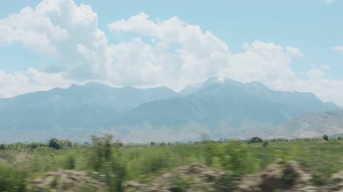 View From the Moving Car on Green Field with Mountains and Beautiful Clouds on a Background in