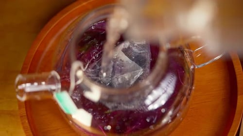 Top view of pouring hot water into transparent glass tea pot with tea bag and herbal leaves. Process
