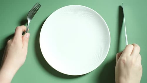 Female hands holding a fork and knife next to an empty plate on a green background close-up top view