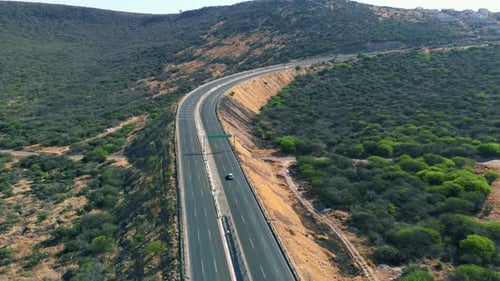 drone view of vehicles driving on a road going up the hill around a green field of trees and morning