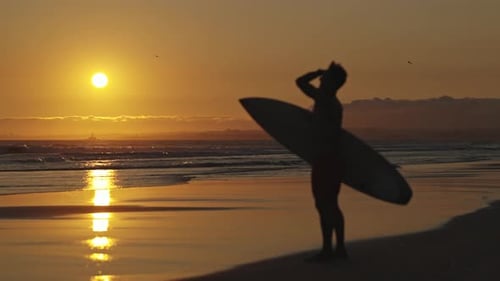 Verschwommene Silhouette eines Surfers, der bei strahlendem Sonnenuntergang mit einem Surfbrett am Ufer steht
