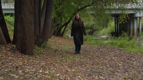 Woman in Dark Coat Wanders Through Forest Near Water Under Cloudy Sky Solitary Woman with Long Hair