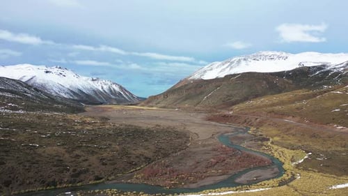 Aerial view drone flying over scenic snowy mountains and a small river with a cloudy sky.