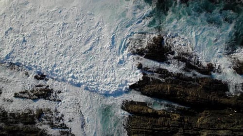 aerial top shot of ocean waves hitting rocky shore crashing violently with sun reflecting on the sur