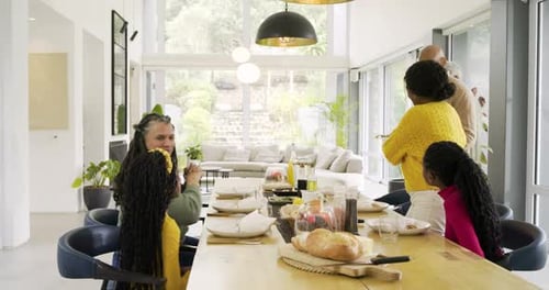 Diverse family greeting arriving grandpa, hugging, passing dishes at dining table to share meal