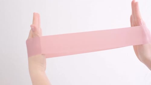 Mãos femininas esticando um elástico rosa de fitness em close-up de fundo branco.