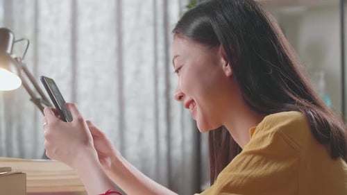 Close Up Of Young Asian Female Studying At Home Smiling And Using Smartphone While Reading Books